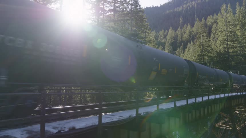 Sunshine filters between tank cars as a crude oil train crosses a trestle over a mountain river