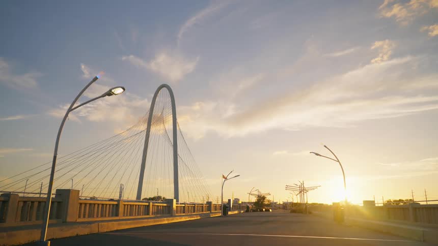 The sun shines brightly upon a bridge as traffic moves on another bridge and the american flag flies in the distance