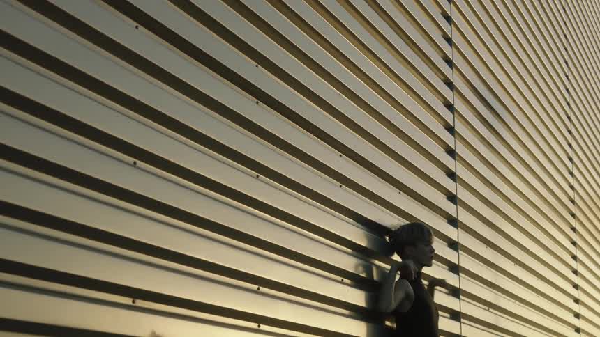 A woman leaning against a building stretches her arms beside her head and rotates her wrists