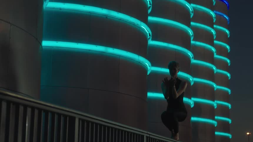 A woman is on a ledge dancing near a building with neon lights glowing in the darkness