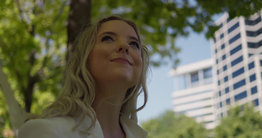 A woman with a camera is under a tree in the city smiling as she looks around