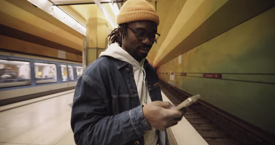 A man wearing a jacket and a knit cap is standing in the subway station texting on his phone as a train passes