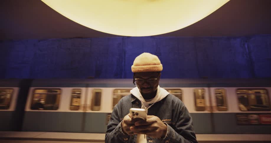 A man wearing a jacket and a knit cap is standing under a light in the subway station texting on his phone as a train passes