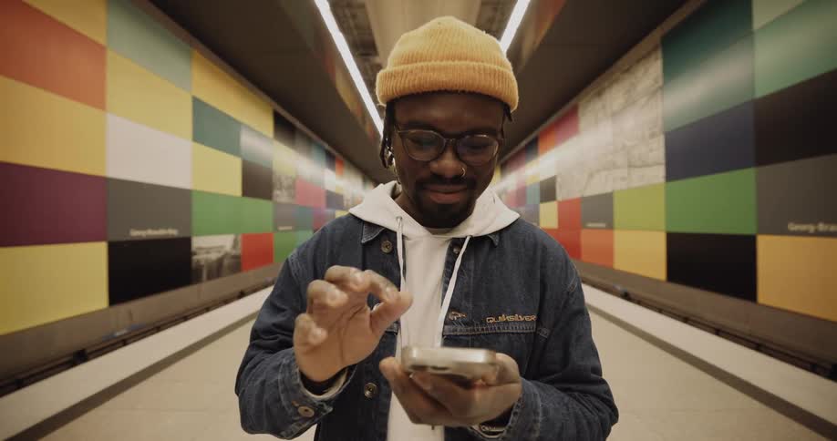 A man wearing a coat and a knit cap is standing in a hallway painted with colored rectangles is holding his phone and pushes a button with his finger