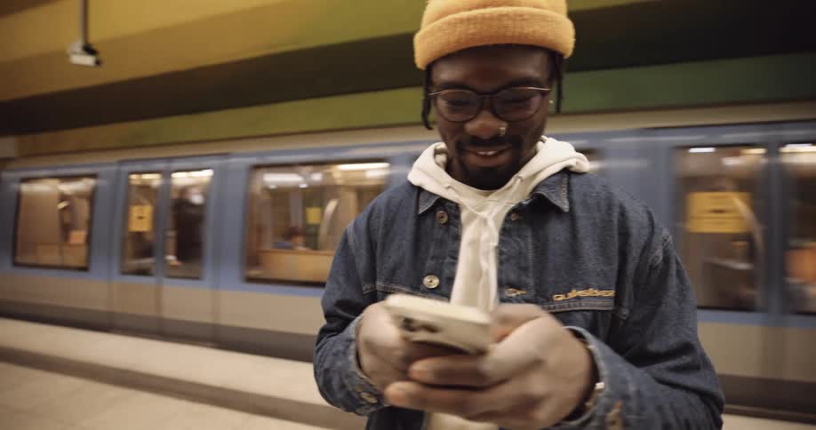 A man wearing a jacket and a knit cap is standing in the subway texting on his phone as a train passes