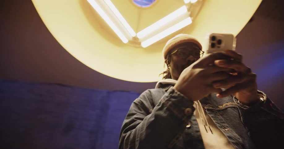 A man standing directly under a light fixture smiling down at his phone as he texts