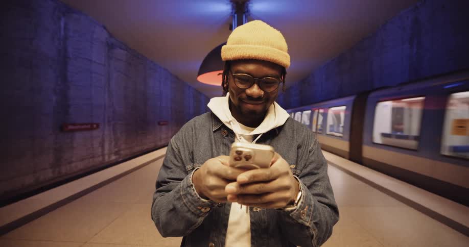A man wearing a jacket and a knit cap is standing in the subway station texting on his phone as a train passes