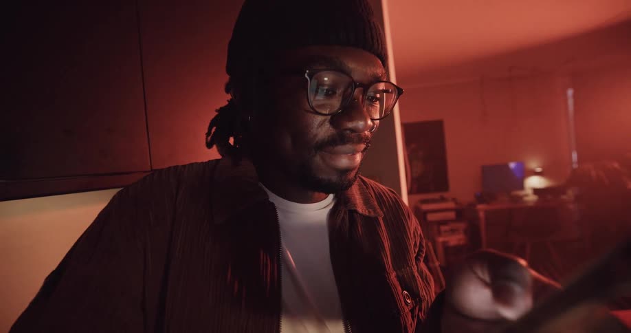A man wearing a knit cap and glasses is standing in a hallway scrolling on his phone and there is a room with a computer on a desk in the distance