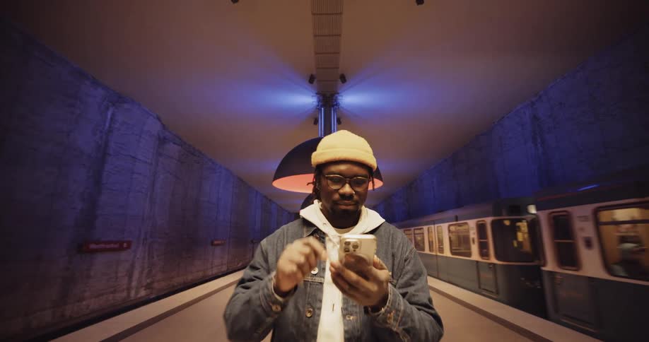 A man wearing a jacket and a knit cap is standing in the subway station texting on his phone as a train passes