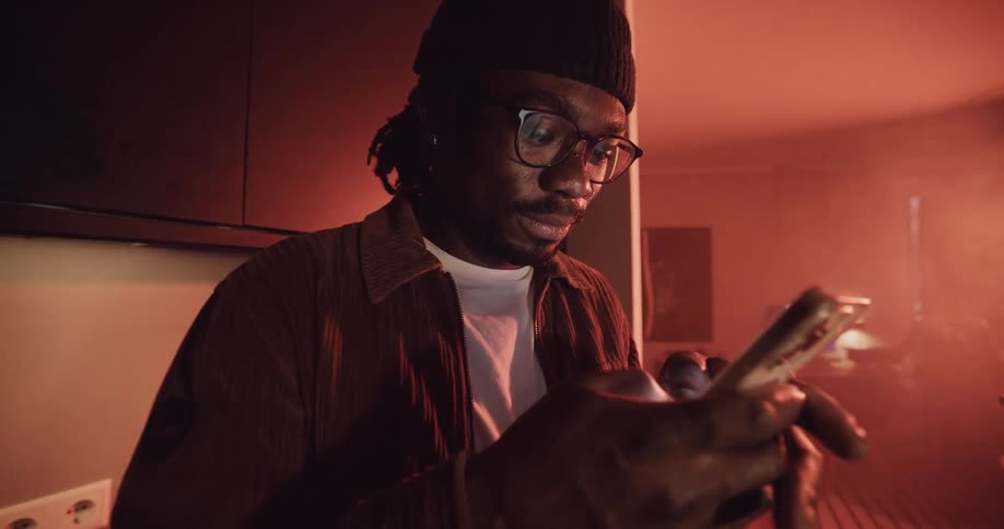 A person wearing ablack hat standing in their kitchen texting on their cellphone