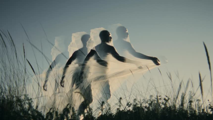 A man is standing outside in the weeds with a sheer cloth over his body as it blows in the wind and ghostly images of him multiply