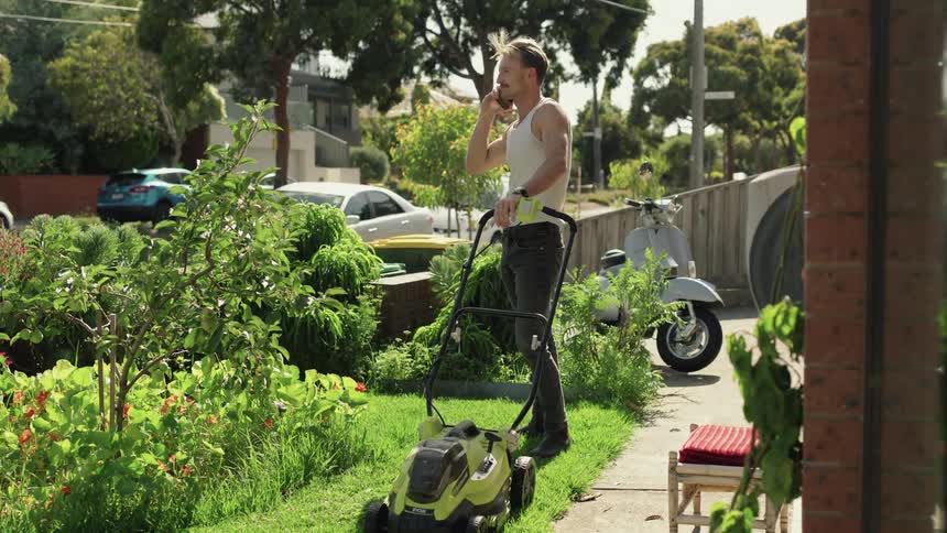 Man using a lawnmower on the yard answers a call on his smartphone