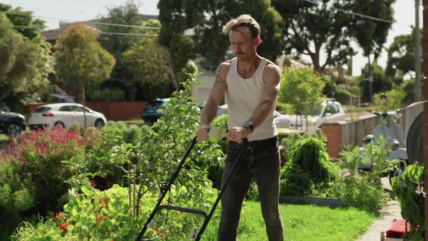 Young man pushes a lawnmower on the front yard of his house at daytime