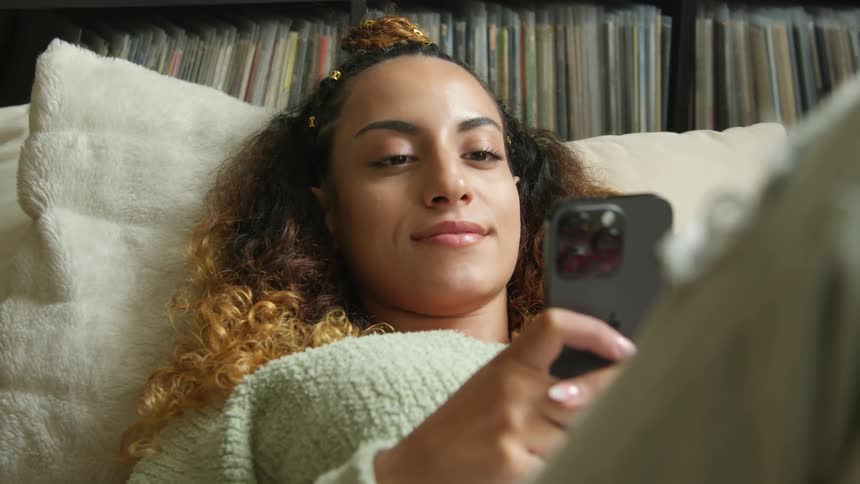 A woman is lying down with her head on a pillow and a smile on her face as she holds her phone