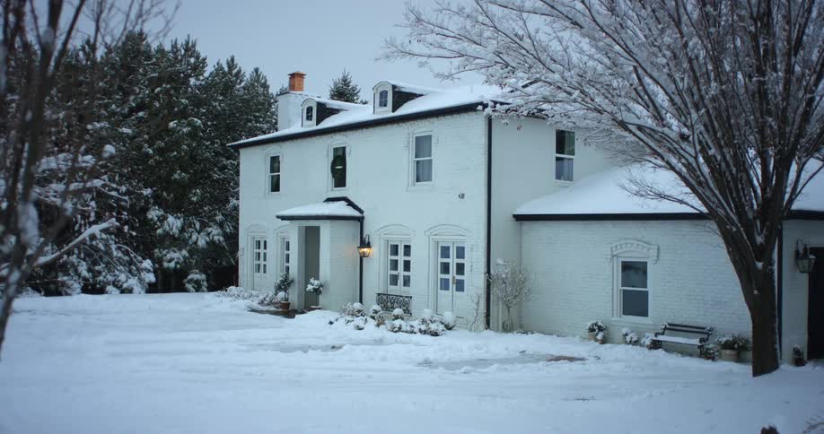 The porch light on a snow covered house is on and the yard is white with snow cover and tree branches are covered with snow