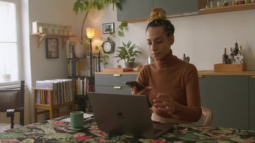 A woman is sitting at a table typing on her laptop computer and she answers her phone