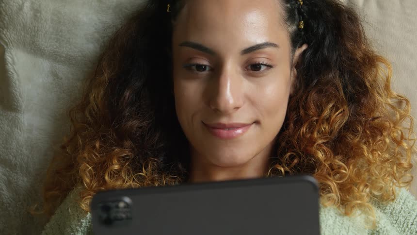 A woman is lying on her bed with her head on a pillow and a smile on her face as she holds a tablet computer