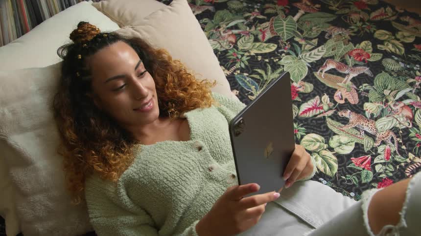 A woman is lying on her bed with her head on a pillow and a smile on her face as she holds a tablet computer