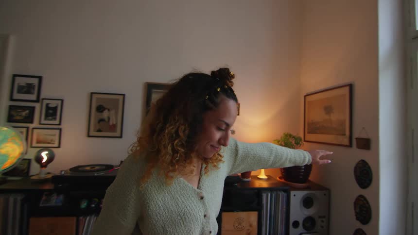 A woman is standing in the living room dancing near her turntable and record collection and turns in front of the windows