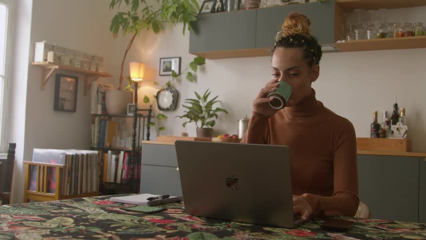 A woman is sitting at a table typing on a laptop computer as she drinks her coffee