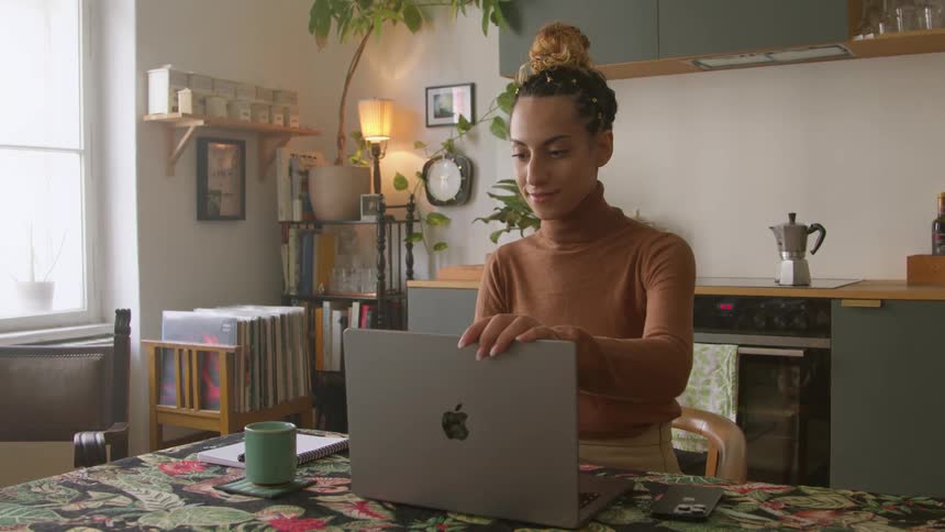 Woman sitting at table near kitchen closes laptop and picks up smartphone
