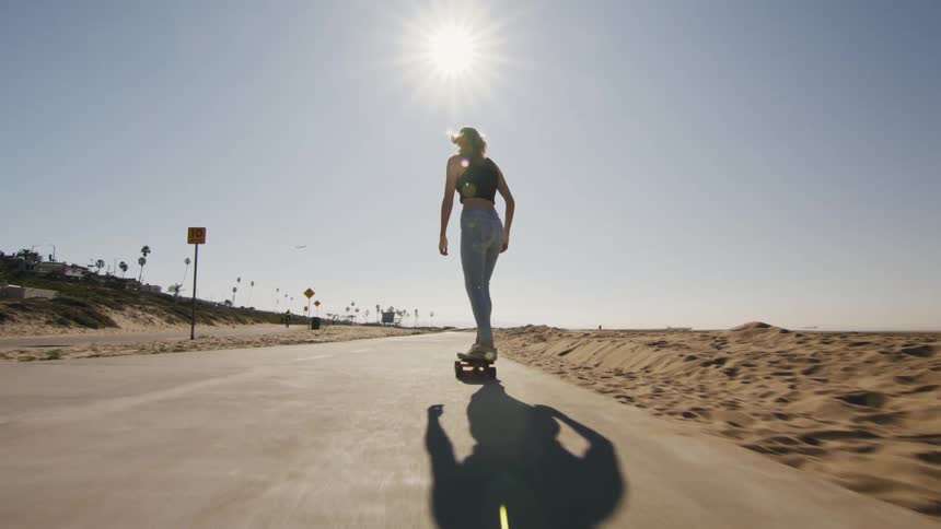 Sun shines over a young fit woman riding a skateboard on a road on a beach at daytime