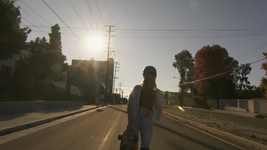 Blonde woman with hardhat silver jacket and jeans holds skateboard and poses for the camera on the street