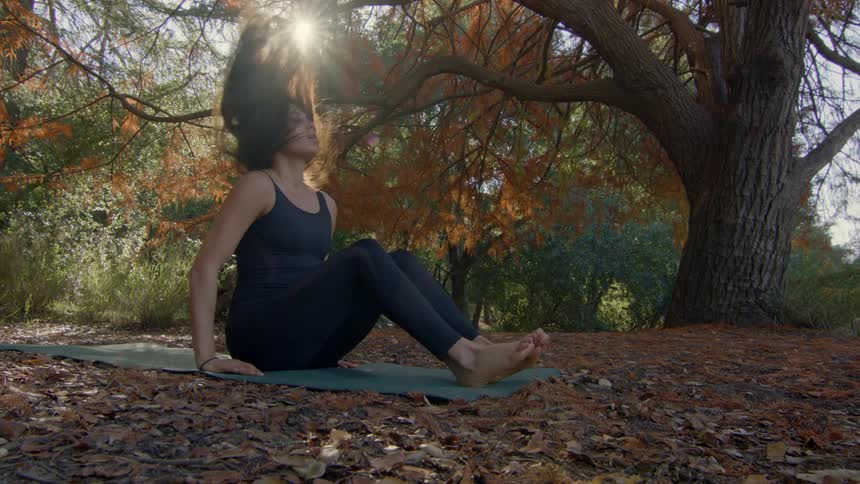 Woman with brown hair black top and leggings stretches on yoga mat in the autumn forest