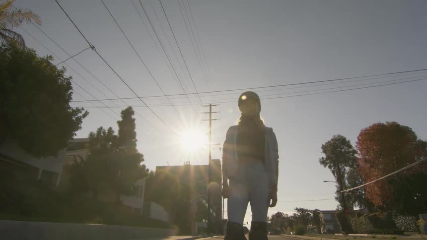 The sun shines brightly upon a teenage girl standing in the street with her foot on her skateboard