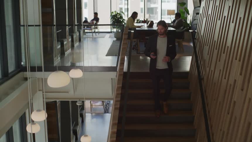 Employees sit at a table in an office as a man looks at his cell phone as he walks downstairs