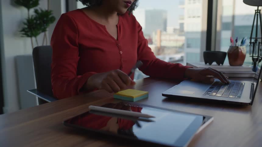 A woman wearing a red shirt typing on a laptop writing something down and then typing again