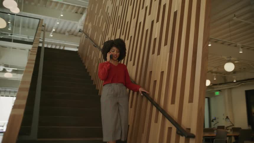Businesswoman with curly hair red blouse and gray pants goes downstairs talking on mobile phone in the office building