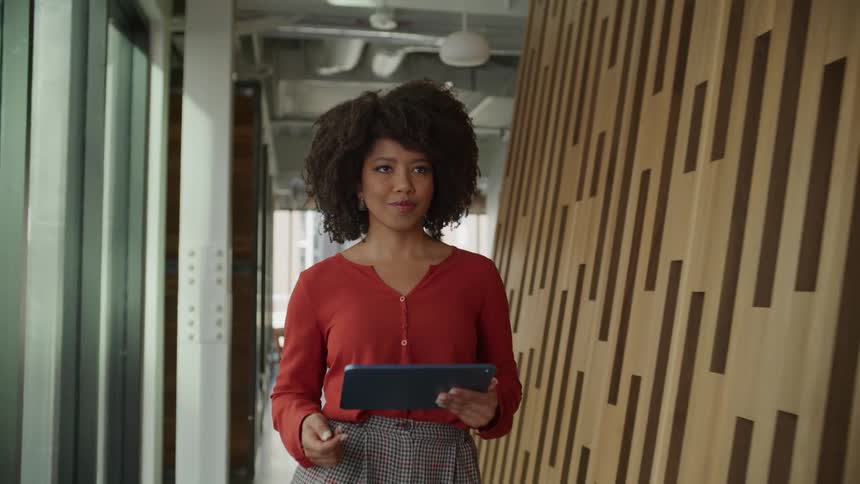 Woman with curly hair and red blouse and gray pants walks corridor using tablet in the office building