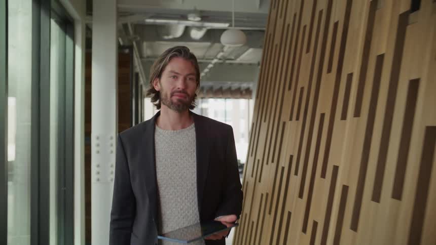 Businessman with long hair beard blazer jacket and brown sweater looks down at tablet walking in the building