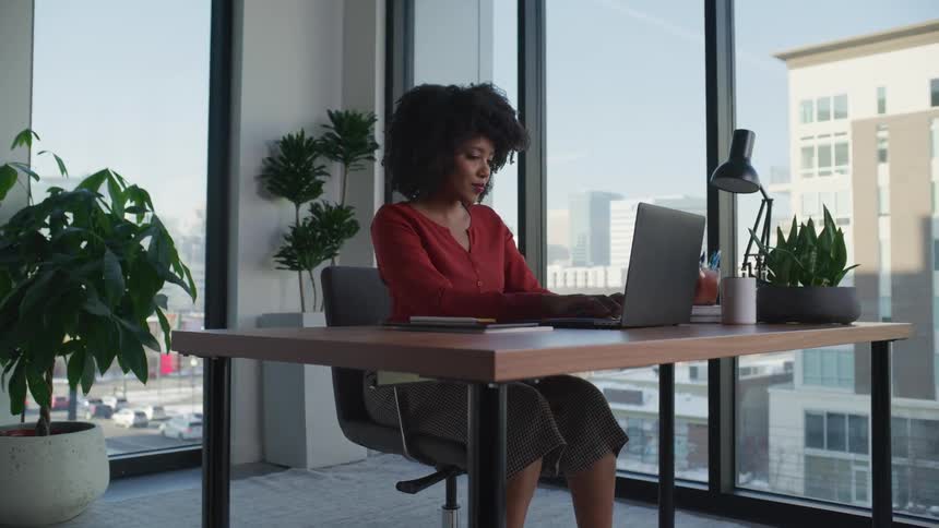 Businesswoman with curly hair red blouse and brown skirt sits at desk working on laptop in the office