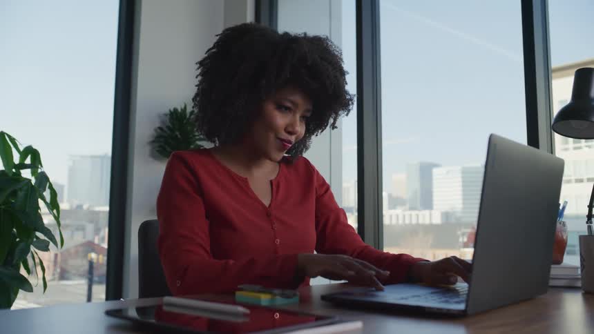 A woman is sitting at a desk in an office working on a laptop computer and taking notes on paper