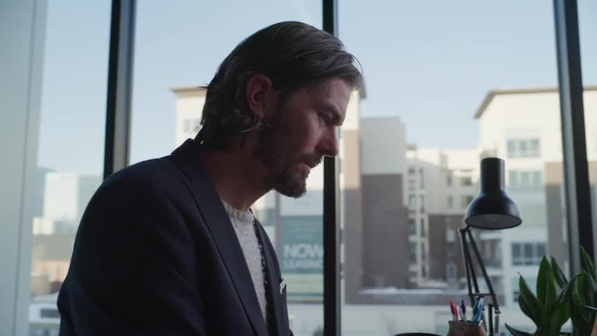 A man wearing a sweater and blazer types on his laptop and then writes something down