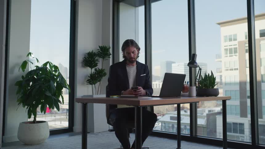 A man sitting at his desk checking his phone and then returning to work at the laptop