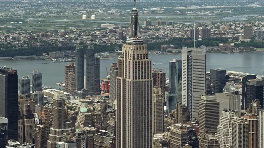 An aerial shot of the iconic Empire State Building in Midtown Manhattan during a sunny summer day with views of Hudson River skyscrapers and surrounding landmarks
