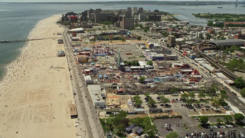 An aerial view of Coney Island beach and boardwalk orbiting Luna Park as sunbathers enjoy the sunshine and amusement park rides