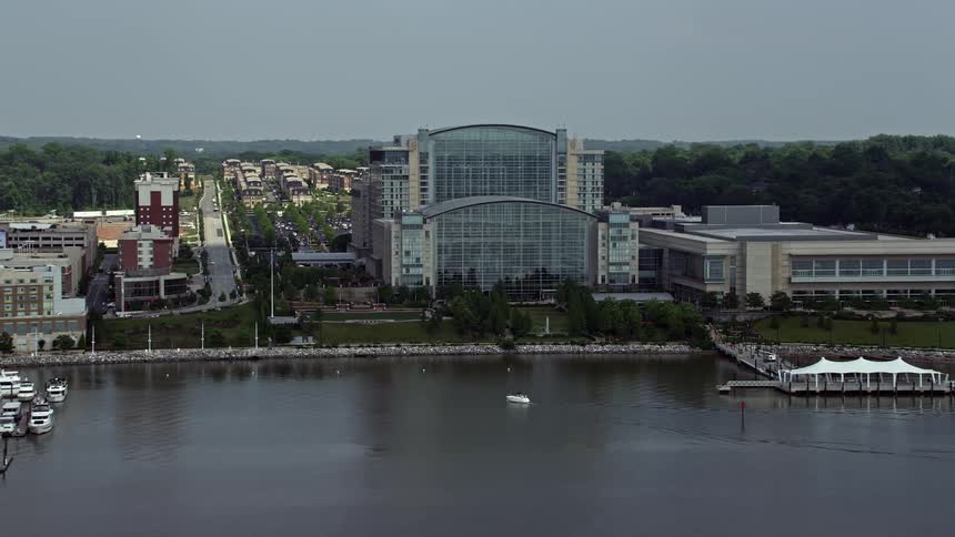 Aerial footage of the Waterfront Gaylord National Resort & Convention Center in National Harbor Maryland on a partly cloudy day with boats floating in the harbor and shoreline visible
