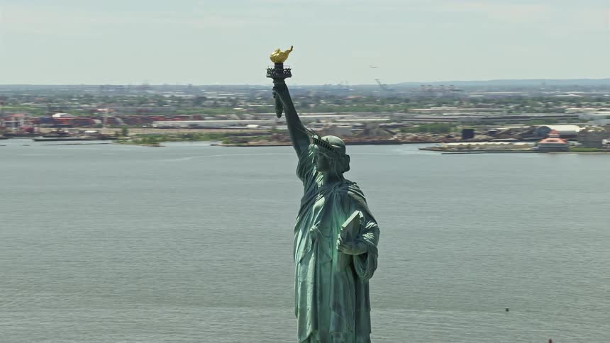 Aerial footage of the Statue of Liberty overlooking New York Harbor on a sunny day surrounded by the city skyline docks and transportation