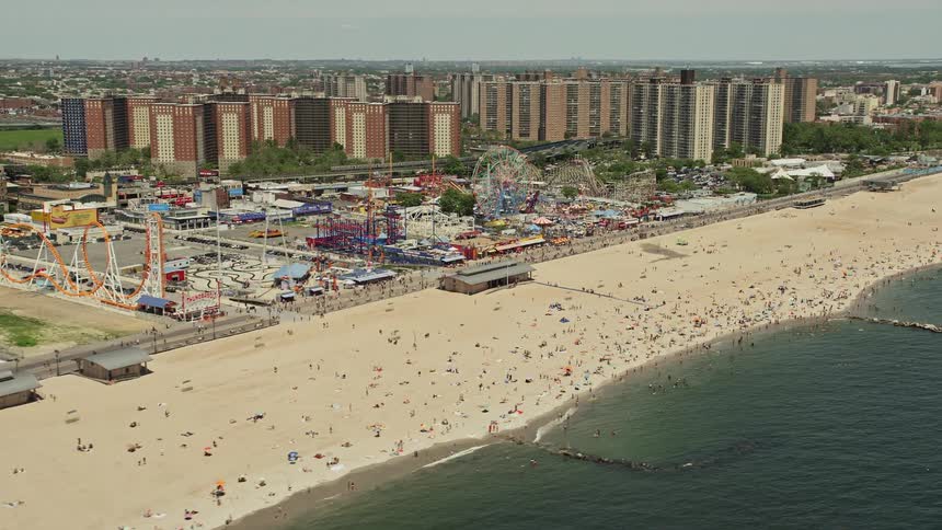 Aerial footage flying over beach goers on Coney Island Beach before approaching the lively Luna Park amusement park on a sunny day in Brooklyn New York City