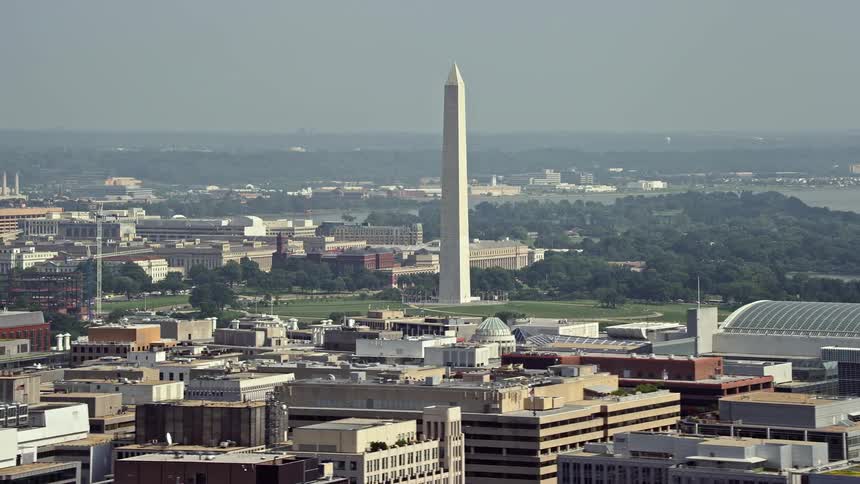 A hazy daytime aerial view of the iconic Washington Monument in Washington DC seen from office building rooftops amidst historical landmarks and tourist attractions