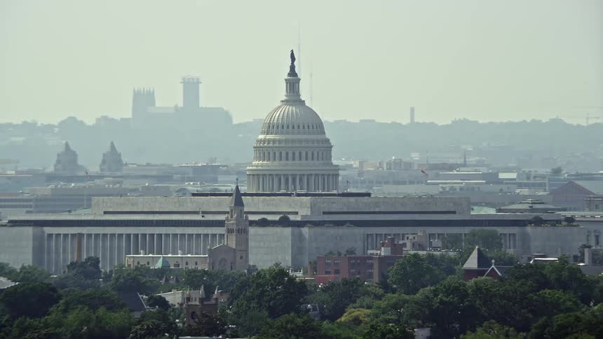 An iconic government building the United States Capitol dome sits behind the Cannon House Offices and James Madison Building in hazy daytime aerial footage of Washington DC during summer