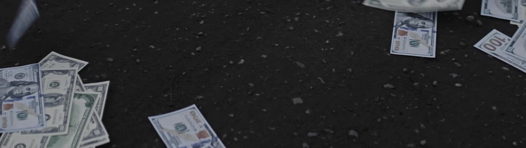 Hundred dollar bills are lying on the ground among pebbles in the dirt and they away in the wind