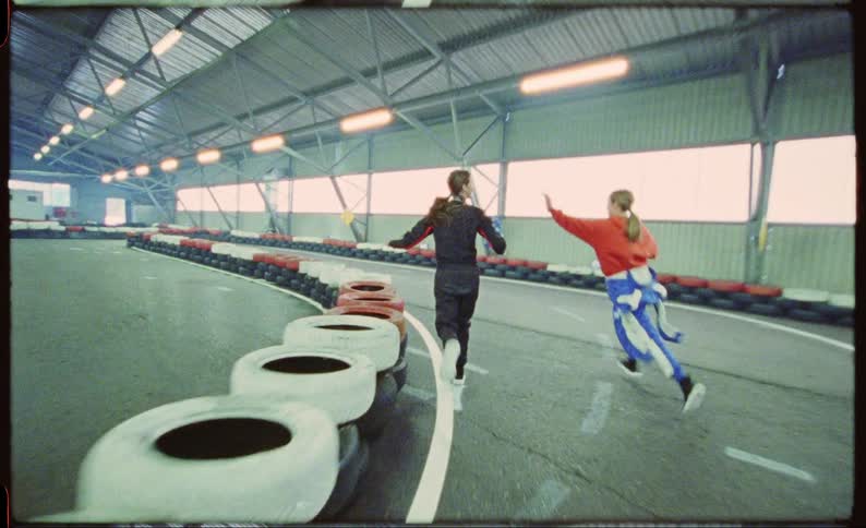 Two girls in racing suits running around a track inside a building