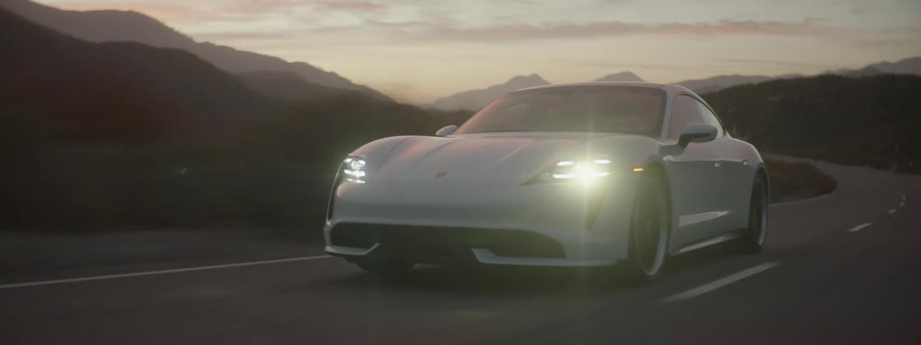 A white sports car with the headlights on is traveling on a desert road with mountains in the distance