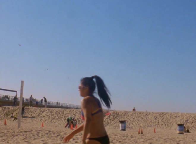 Woman wearing bikini serves volleyball on sand as people move around in background