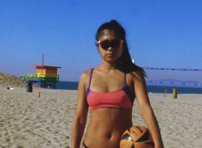 A teenage girl wearing sunglasses and a swimsuit is standing on the beach holding a ball